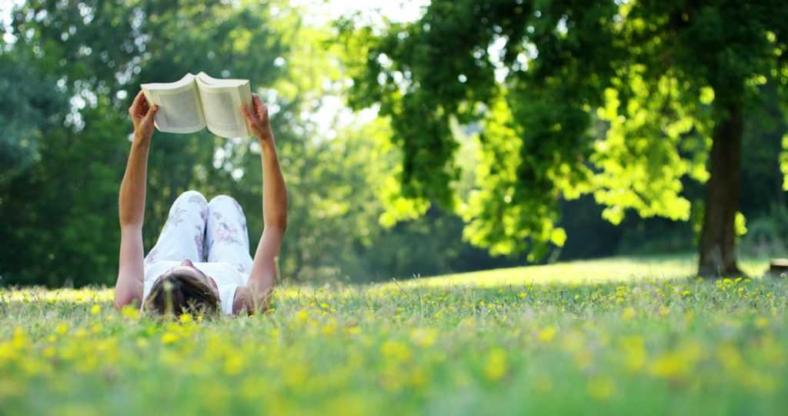 Leżąca kobieta na trawie, czytająca książkę trzymając ją nad głową